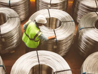 Overhead: Industrial worker (white hard hat, yellow-green vest) examines a large spool of metal wire/rebar in a warehouse.