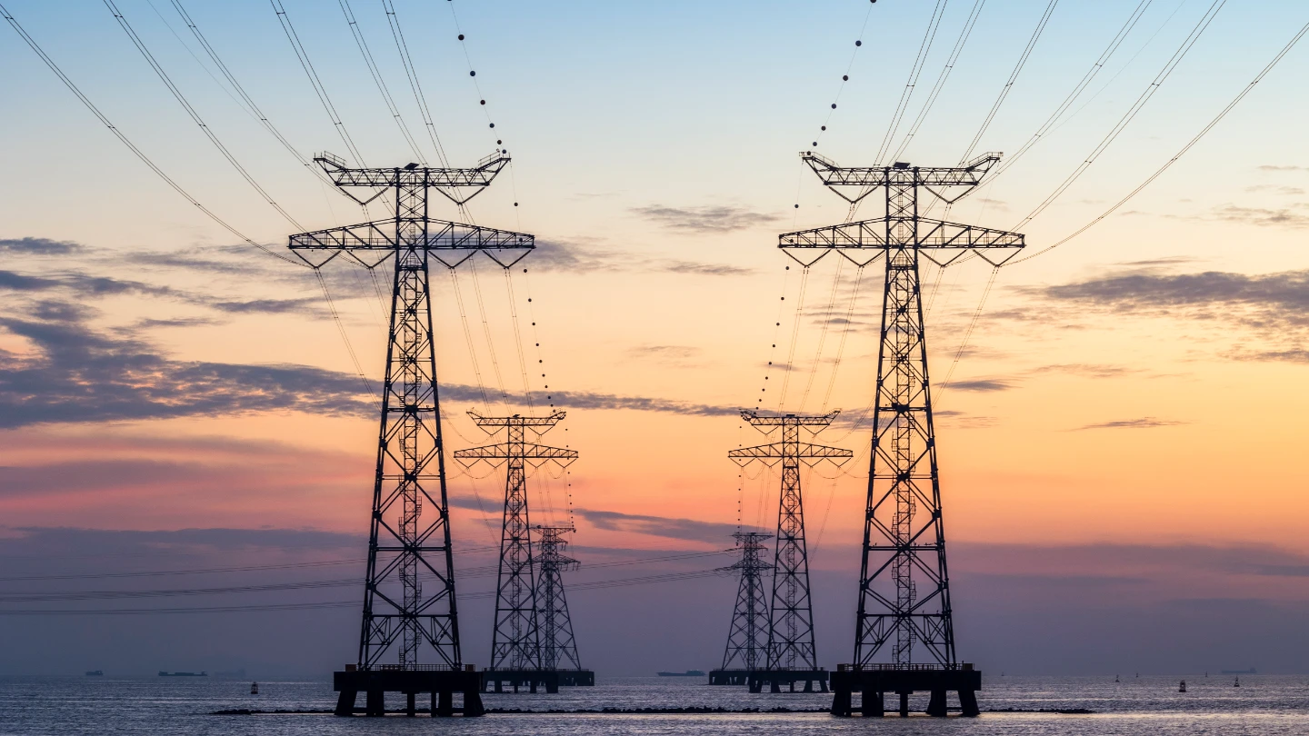 Large metal pylons carry power lines over water, silhouetted against a dramatic sunset of blue to orange.