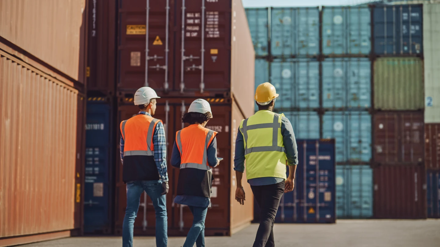 Two men & one woman in hard hats/safety vests walk away through a container terminal, with tall stacks of red/blue shipping containers behind.